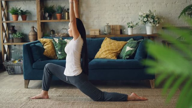 Female Asian gymnast is training at home practising splitting legs sitting on carpet in living room. Active lifestyle, flexibility and sporty young people concept.