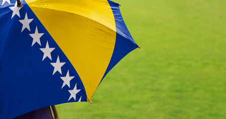 Bosnia and Herzegovina flag umbrella. Close up of printed umbrella over green grass lawn / field. Rainy weather forecast concept.