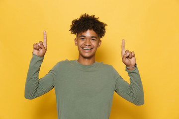 Portrait of joyous african guy having stylish afro hairdo pointing fingers upward at copyspace, isolated over yellow background
