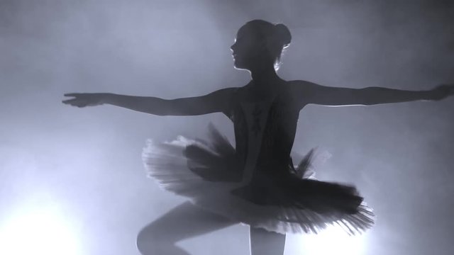Ballerina's silhouette turning around. ballerina with hair in a bun is dancing on a scene, against bright light of soffit and smokescreen