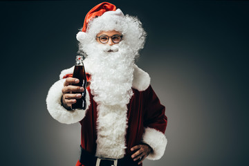 cheerful santa claus in costume showing cream soda bottle isolated on grey background