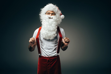 Fototapeta na wymiar happy santa claus in eyeglasses holding suspenders standing isolated on grey background