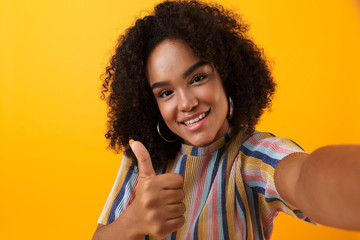 Young happy african cute girl posing isolated over yellow background take a selfie with thumbs up gesture.