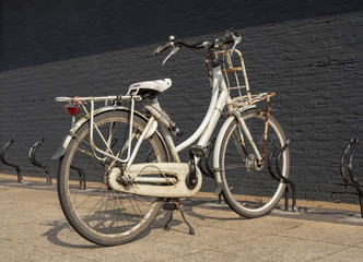 Fototapeta na wymiar Old rusty grunge vintage white bike in bicycle rack in front of a black painted brick wall