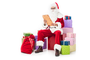 santa claus in eyeglasses reading old parchment and sitting on pile of gift boxes isolated on white background