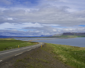 Asphalt road line through rural north summer landscape with green grass. colorful cliffs, lake, blue mountains and dramatic sky, Iceland western fjords, golden hour light