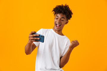 Young african man posing isolated over yellow background play games by phone make winner gesture.