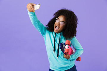 African girl posing isolated over violet background holding skateboard take a selfie by phone.