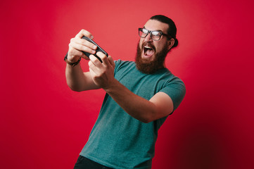 Image of amazed bearded man playing on smartphone over red background