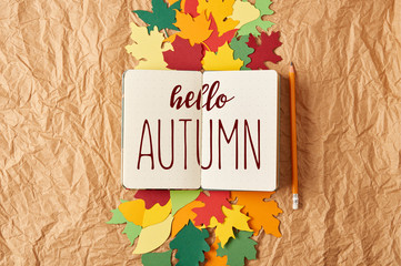 flat lay with "hello autumn" inspiration in notebook, pencil and colorful handcrafted paper leaves on crumpled paper background