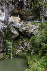 Spain, Asturias, Picos de Europa, Covadonga: View of The Holy Cave of Covadonga (Santa Cueva) a famous Spanish sanctuary with Hermitage, waterfall and small water reservoir in rocky mountains.