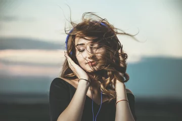 Poster romantic portrait of a beautiful girl in headphones with flying hair from the wind, young woman listening to music outdoors © fantom_rd