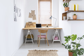 Real photo of a simple home office interior in a bright room with a desk, window blinds and plant