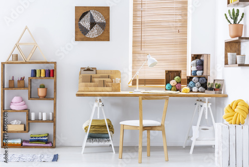 Real photo of a creative room interior with a desk, threads, knitting wool and graphic on a wall