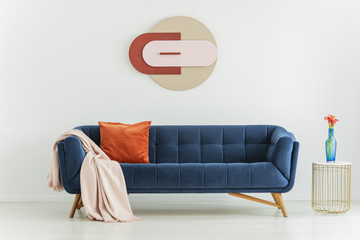 Flower on table next to blue couch with orange pillow and blanket in interior with poster. Real photo