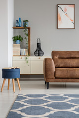 Navy blue stool next to leather sofa in grey living room interior with poster and carpet. Real photo