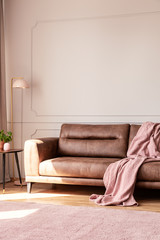 Pink blanket on leather couch in white living room interior with lamp and plant on table. Real photo