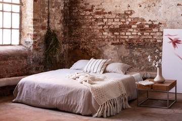 Bed with blanket and cushions next to table with flowers in bedroom interior with window. Real photo