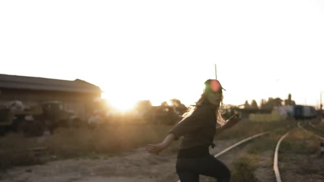 Long haired woman in cap running away from a creepy zombie. Male and female zombie chasing girl going by railway with an abandoned town on the background