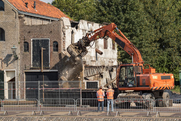 Dismantling of an old house with an excavator while two workmen supervising the demolition