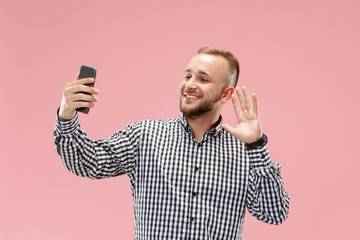 Portrait of attractive smiling young man taking a selfie with his smartphone. Isolated on pink studio background. Human emotions, facial expression concept. Trendy colors