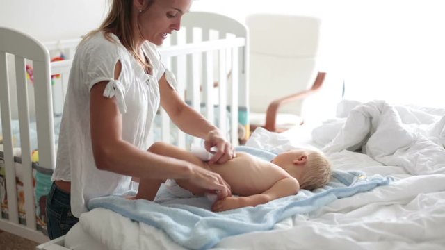 Young mom, changing baby diaper after bath in sunny bedroom