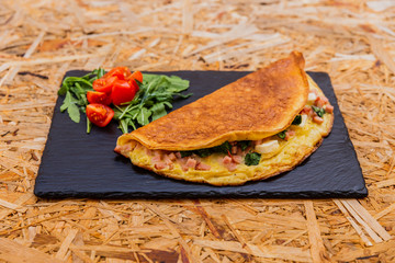 A horizontal photo of omelette with ham, spinach and cheese, garnished with rocket salad and cherry tomatoes. Served on a black stone plate.