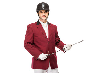 smiling young horseman in uniform holding horseman stick and looking at camera isolated on white