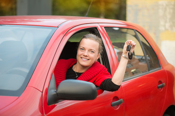 A beautiful young girl dressed in a red coat sits behind the wheel of a red car, smiles and holds car keys