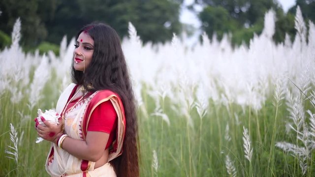 A beautiful and religious married Indian woman holding a shankha or conch shell looks behind in a field of white flowered grass or kaash phool