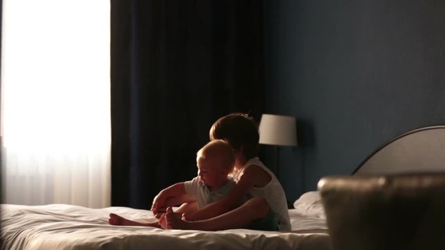 Two sibling children, baby boy and his older brother, sitting on bed in sunny bedroom, playing together, back light
