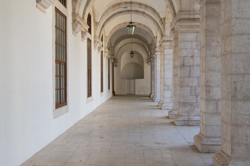 Fototapeta premium Long passageway moving to perspective under arches with stone block columns on one side and a white wall on the other side in the cloisters at the Convento da Graca in Lisbon, Portugal