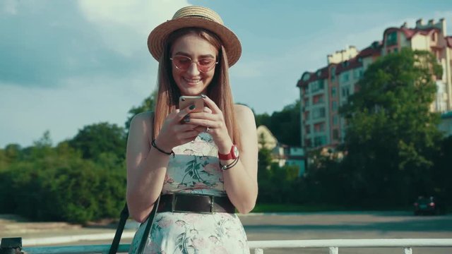 Attractive young woman in trendy summer look actively uses her phone via internet browsing outdoors, texting and smiling to phone screen. Social networks, communication. Female portrait
