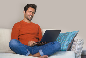 young happy and attractive man working relaxed with laptop computer at modern apartment living room sitting at sofa couch typing and networking confident and positive