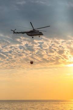 A fire helicopter collects water into the sea and flies toward the mountains to extinguish a forest fire on the background of a beautiful dawn sky. Rescue operation. Forest Fire Prevention