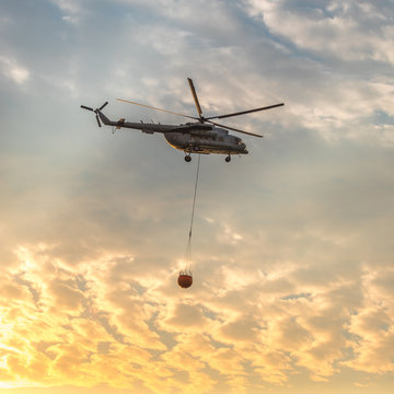 A fire fighter helicopter with a full basket of water flies against a beautiful sunset sky. Rescue operation. Forest Fire Prevention