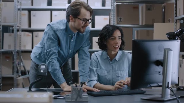 Male and Female Warehouse Inventory Managers Talking, Solving Problems, Using Personal Computer and Checking Stock. In the Background Rows of Shelves Full of Cardboard Box Packages.
