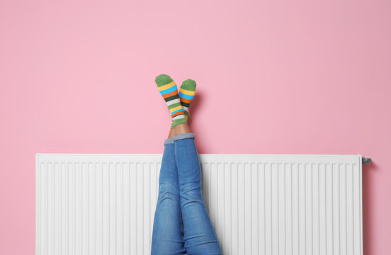 Woman warming legs on heating radiator near color wall