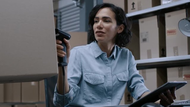 Female Inventory Manager Scans Cardboard Box and Digital Tablet with Barcode Scanner, Worker Puts Package on the Designated Shelf. In the Background Rows of Cardboard Boxes