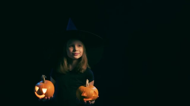Happy joyful girl wearing black witch hat holding jack-o'-lanterns dancing looking out of frame, over black background