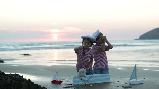 Sailor baby boy, cute child, playing on the beach with wooden boat, fishes and fishing rod on sunset by the ocean