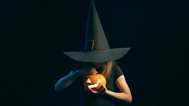 Girl wearing black witch hat holding jack-o'-lanterns opening a pumpking and making a scary face, over black background