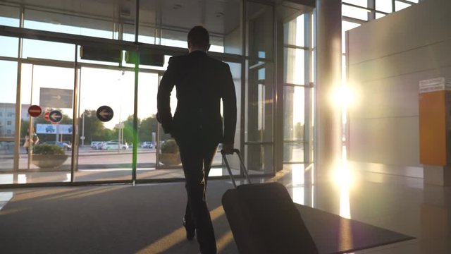 Unrecognizable man walking through glass automatic door of modern airport to city street and pulling suitcase on wheels. Businessman going from terminal to cars parking with his luggage. Rear view