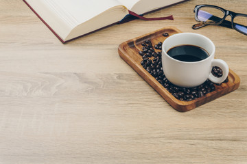 a cup of coffee and a pile of coffee beans in the wooden tray, a red book and an eyeglasses on the wooden table.