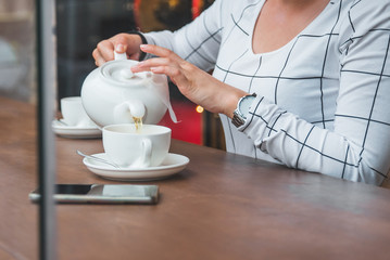 woman pouring tea from white kettle to cap in cafe
