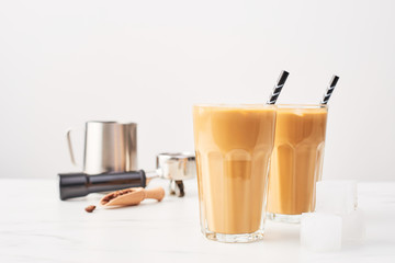 Barista concept. Ice coffee in a tall glass with straws and milk pitcher, portafilter and wooden coffee measure on white marble table over white background. Selective focus and copy space.