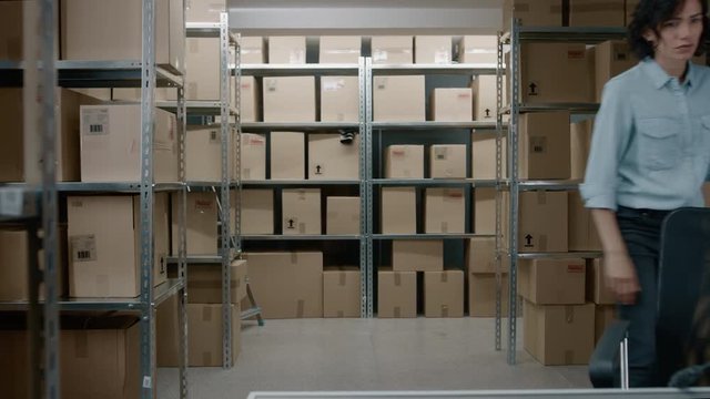 Inventory Management Team Works in a Warehouse Taking Stock, Checking Cardboxes on Shelves, Woman Verifies with Order List. In the Background Rows of Shelves with Filled with Parcels.