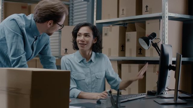 Male and Female Warehouse Inventory Managers Talking, Using Personal Computer and Checking Stock. In the Background Rows of Shelves Full of Cardboard Box Packages.