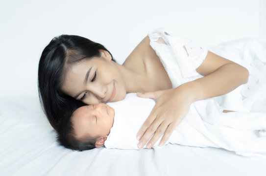 Young Asian mother kissing newborn baby sleeping in the bed, maternity concept, soft image of beautiful family, mother's day concept.
