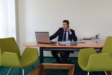 Young businessman wearing suit sitting alone at office desk talking on phone and working with laptop. 